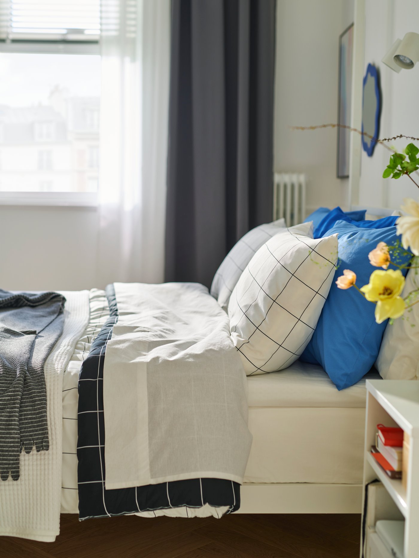 View of bed from the side with white bedding and blue pillows. Window in the background, with dark grey curtain at the side.