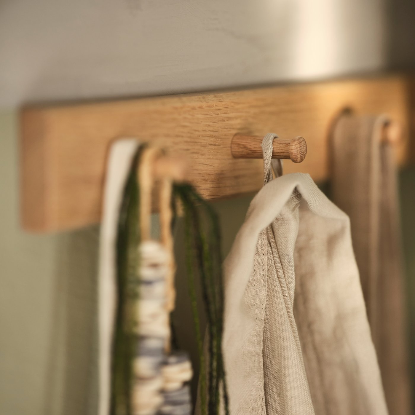 An oak HÖVOLM rack with 6 knobs on a wall, with necklaces and beige fabric hanging from it.