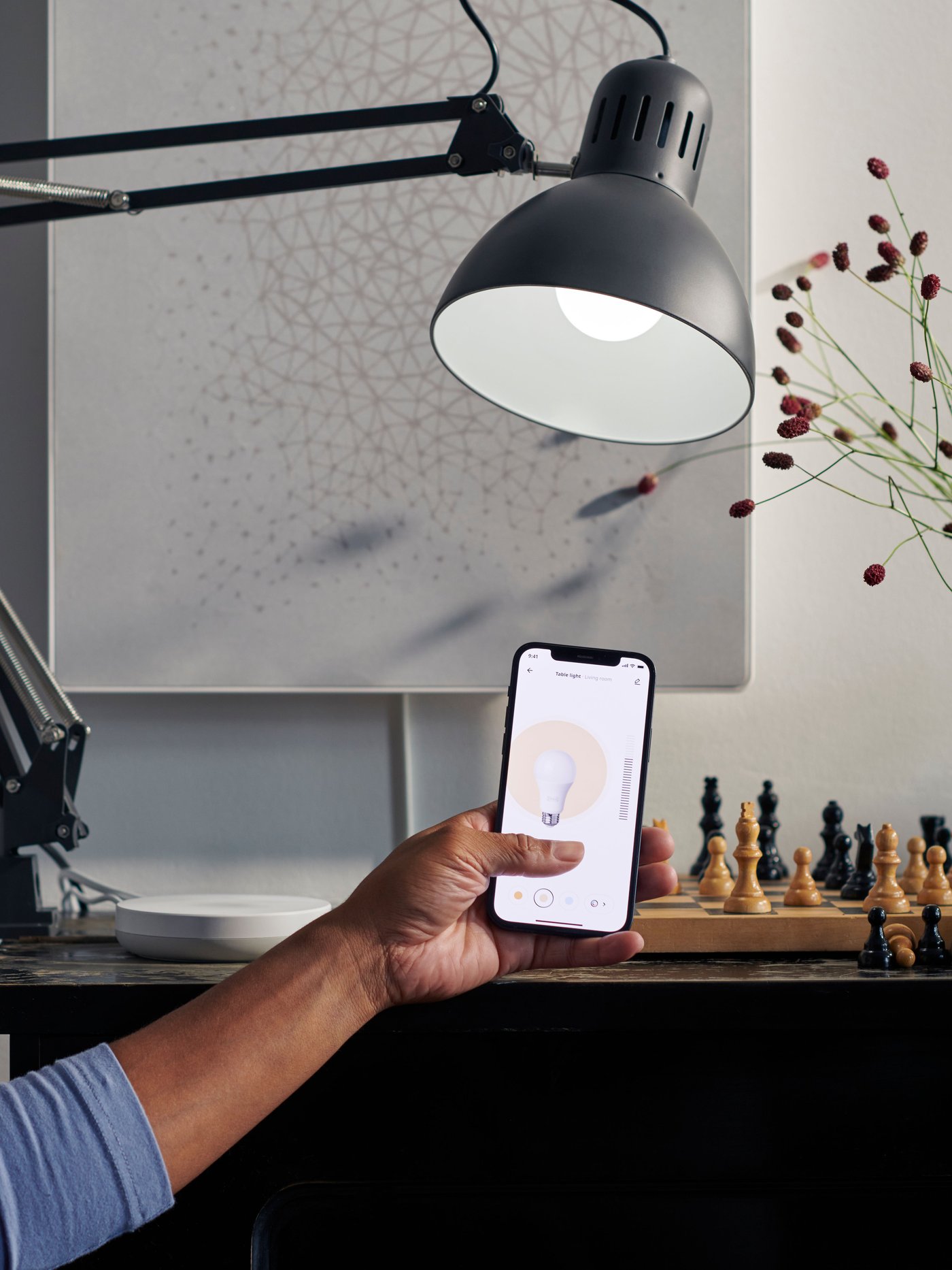A person controls a lamp via the IKEA Home Smart app on a smart phone. A DIRIGERA hub is displayed on a table behind.