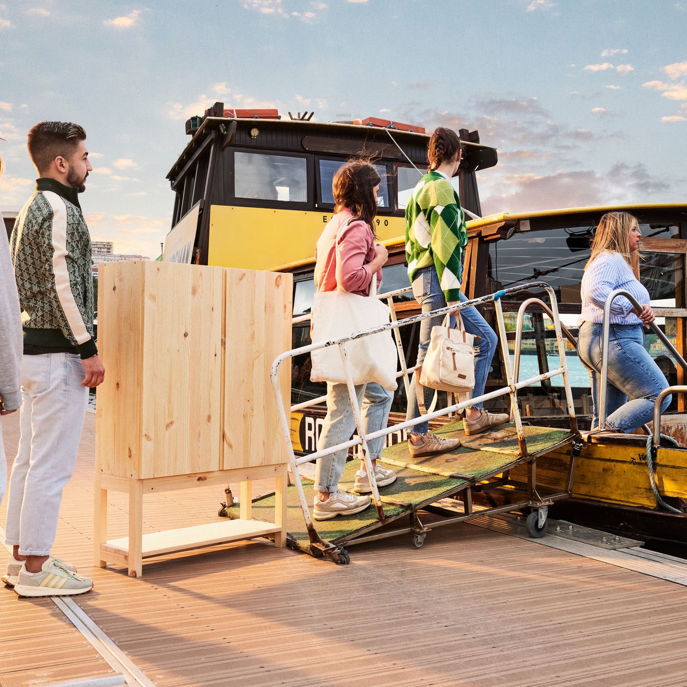 Personer med olika möbler går ombord på ett gulsvart fordon via en ramp, med himmel i solnedgång i bakgrunden.