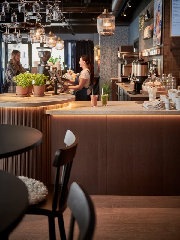 Modern kaffebar med två vuxna, växter och koppar på disken, kaffemaskiner i bakgrunden och varm belysning ovanför.