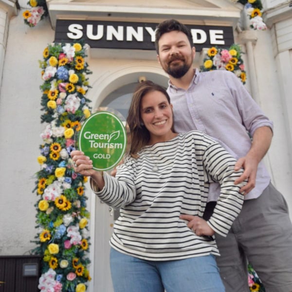 The couple of Sunnyside standing in front of their guesthouse holding a green, round sign that says Green tourism Gold.