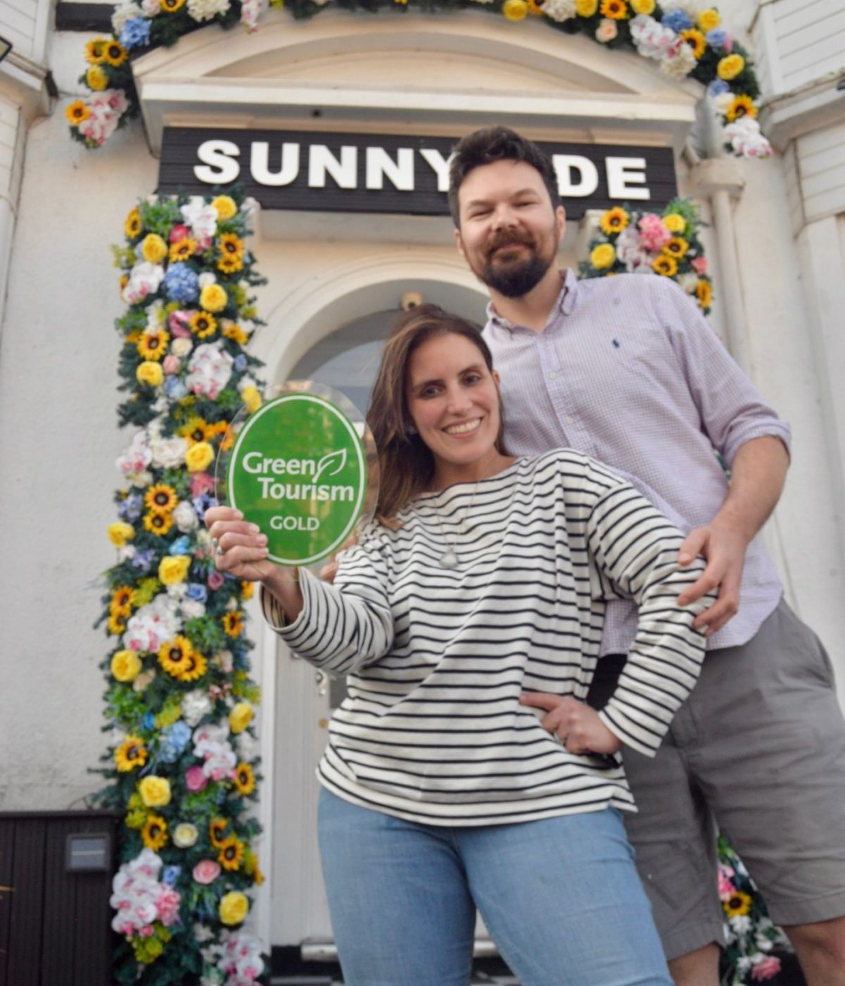 The couple of Sunnyside standing in front of their guesthouse holding a green, round sign that says Green tourism Gold.
