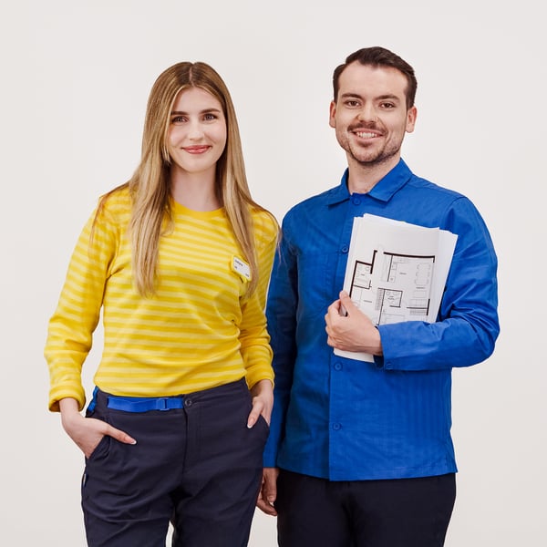 Projektant IKEA stojący w niebieskiej koszulce z projektami w ręku. Obok niego stoi konsultantka IKEA w żółtej koszulce.