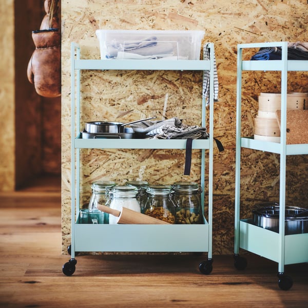 Two green NISSAFORS trolleys are filled with cookware stored in a SAMLA box, KONCIS tins, KLOCKREN steamers, ingredients in transparent KORKEN jars, and other kitchen accessories.