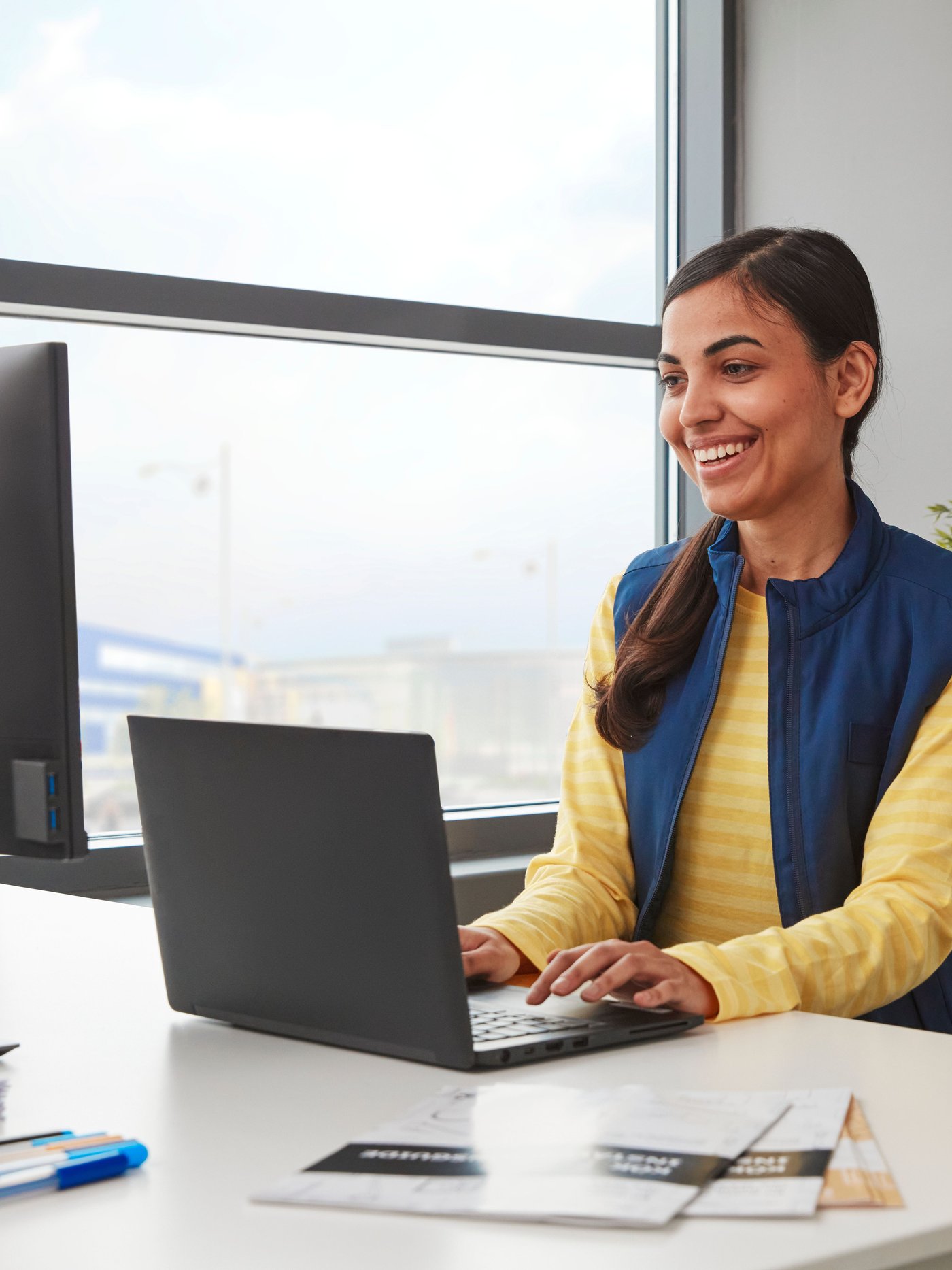 A smiling person in a yellow sweater and a blue vest is working on a black laptop placed on a white desk next to a window.