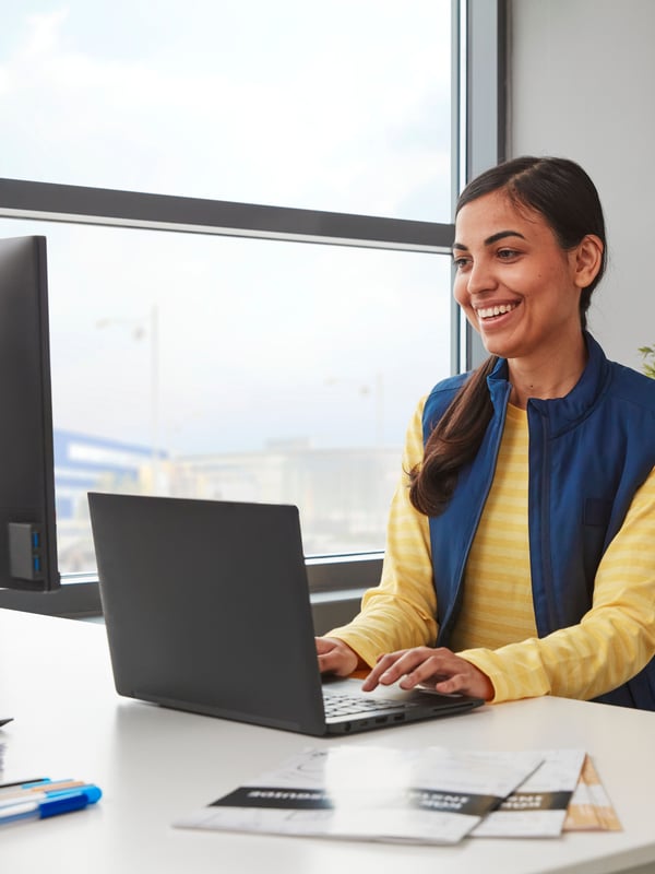 A smiling person in a yellow sweater and a blue vest is working on a black laptop placed on a white desk next to a window.