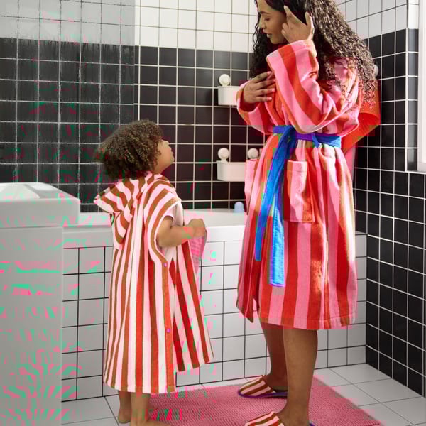 A woman wearing SKOGSSALLAT bathrobe with pink and red stripes and a child in a red striped poncho. Black-and-white bathroom.