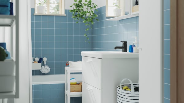 A blue-tiled bathroom with a bathtub has a white ENHET base cabinet, a wall cabinet with mirror doors and a VESKEN trolley.