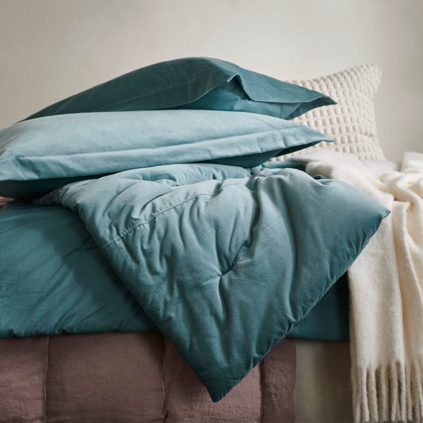 Close up of dark grey-turquoise SANELA comfoter and pillows and a white KLIPPOXEL throw on top of a brown textile.