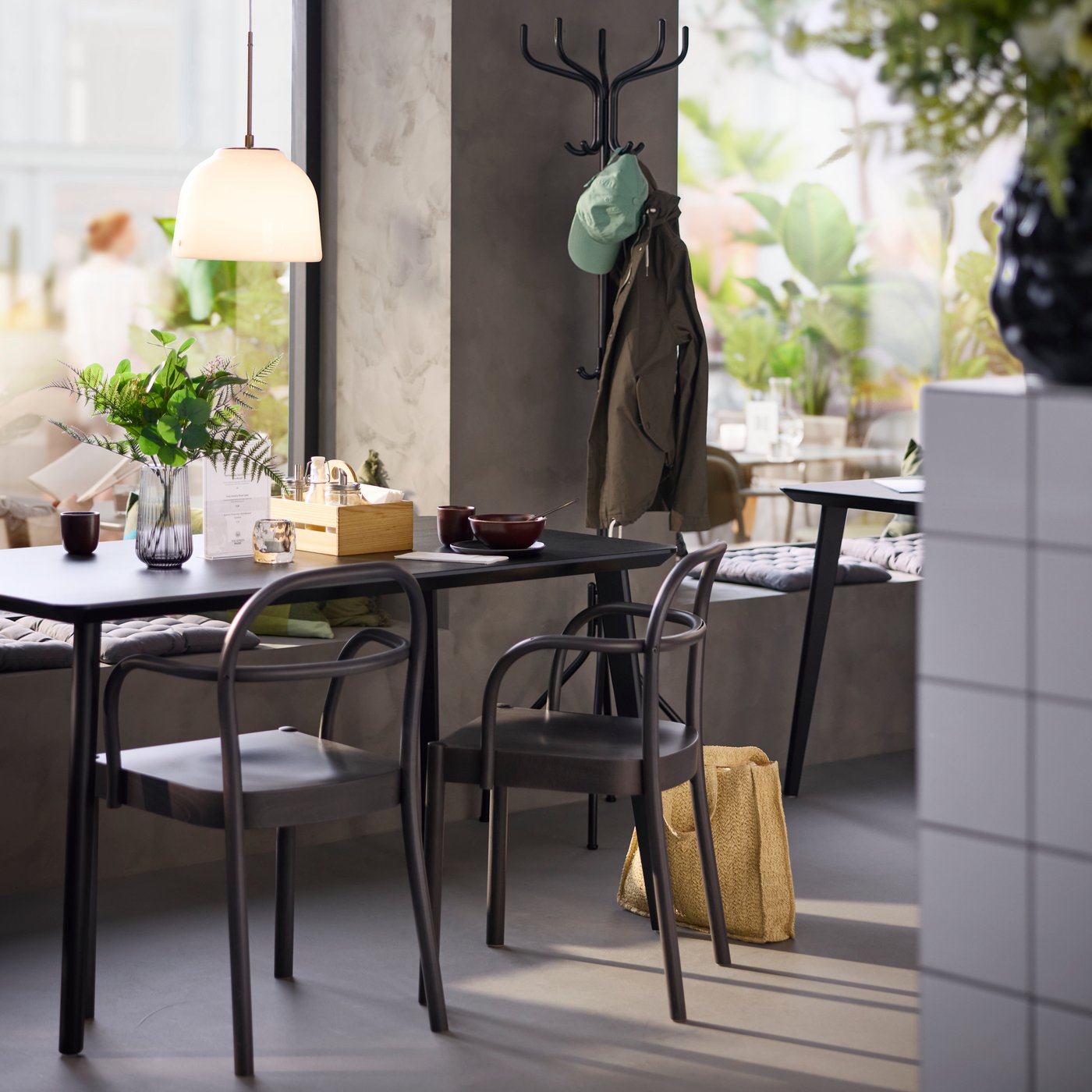 Café scene with natural light, dark brown STOCKHOLM 2025 chairs by a table, with a hat and coat stand in the background.