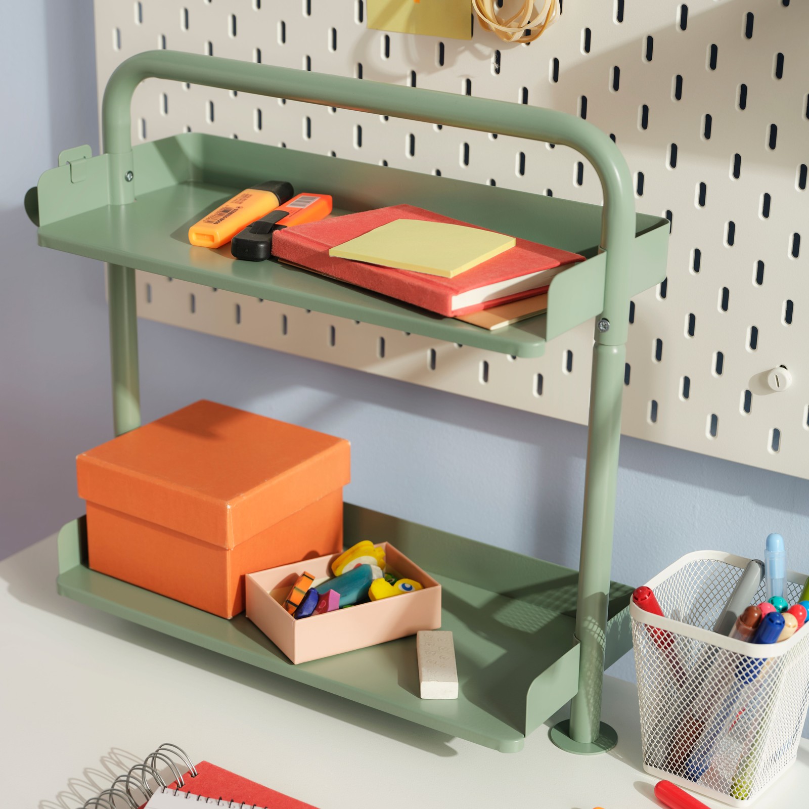 A light grey-green RELATERA desktop shelf on a white desk with stationery on it, and a white SKÅDIS pegboard behind it.