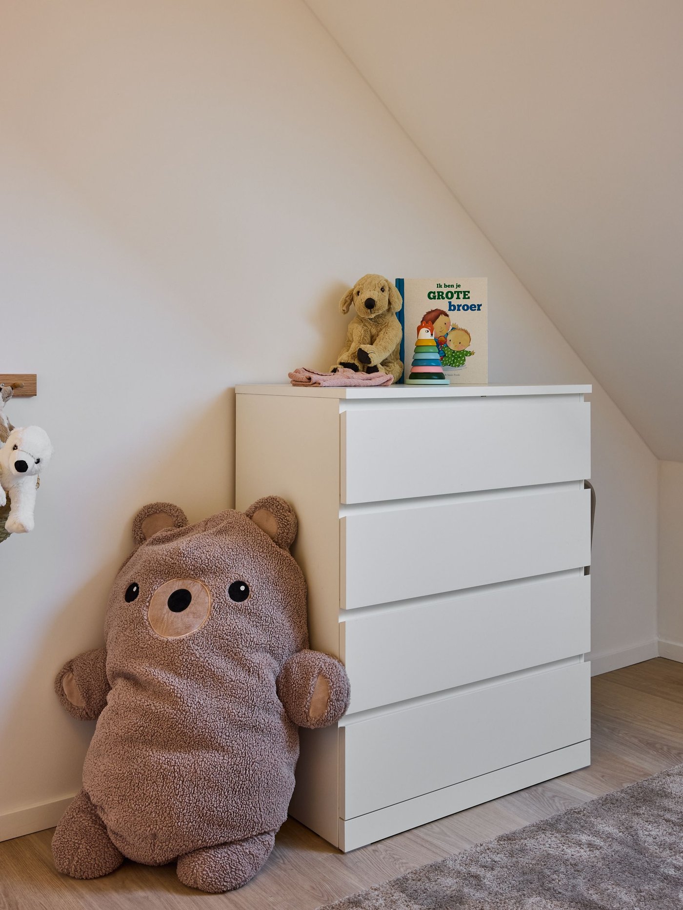 Kinderkamer met witte commode, knuffels en schuin dak, zachte kleuren en warme, rustige sfeer.