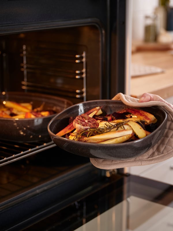 Una mano guantata da forno estrae una teglia ovale GLADELIG grigio scuro piena di verdure.