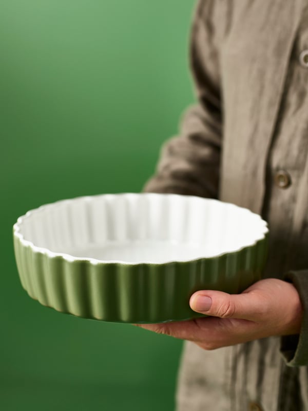 A person wearing a dark beige shirt is holding a white/dark green NÄBBFISK pie dish in a space with green walls.