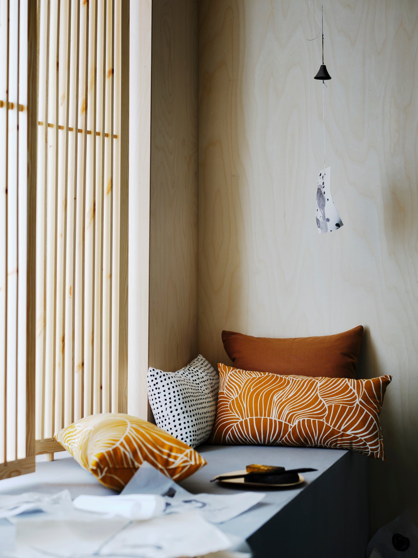 A dark yellow SVAMPMAL cushion with a white mushroom print is on a bench by a window with other cushions.