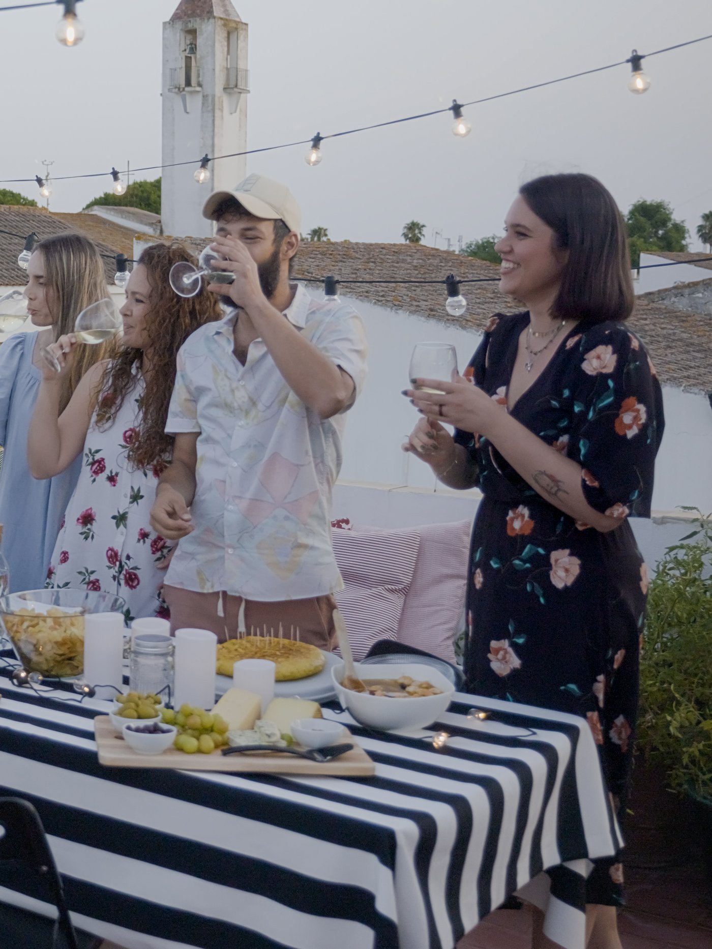 Un groupe de jeunes, debout devant une table de buffet pleine de nourriture sur une terrasse extérieure, mangent, boivent et discutent dans la joie et la bonne humeur.