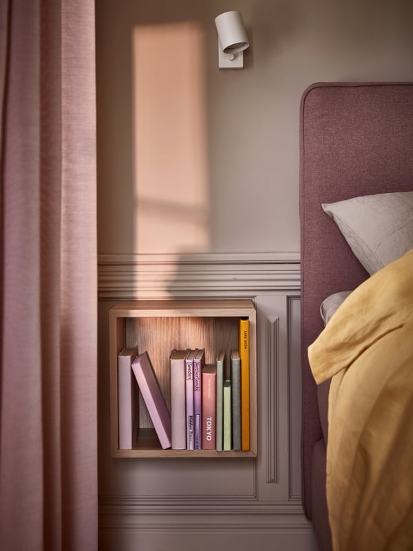 A cube-shaped shelving unit containing books is hung on a wall decorated with wooden pallets in turn of the century style next to a pink box spring bed.