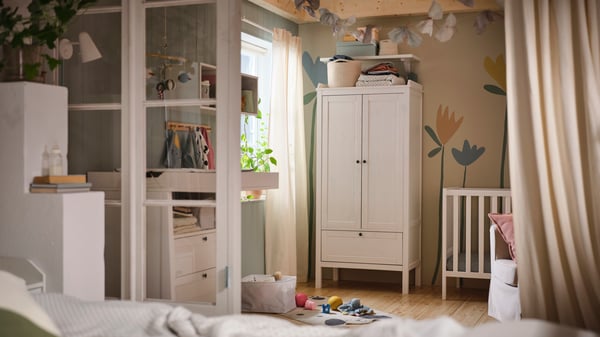 A white SUNDVIK wardrobe against a wall that has a flower mural on it in a baby’s bedroom with a white SUNDVIK cot beside it.