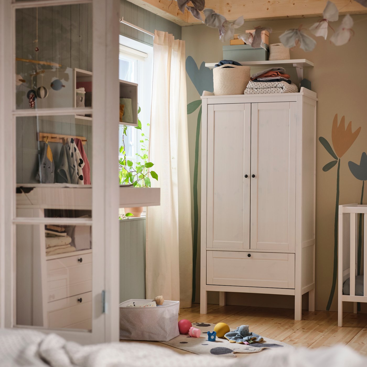 En vit SUNDVIK garderob står mot en vägg med en blommig väggmålning i ett babyrum, med en vit SUNDVIK spjälsäng intill.
