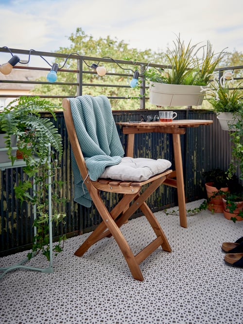 A balcony prepped for spring with light grey ALTAPPEN floor decking, plants, a colourful light chain, table and chair.