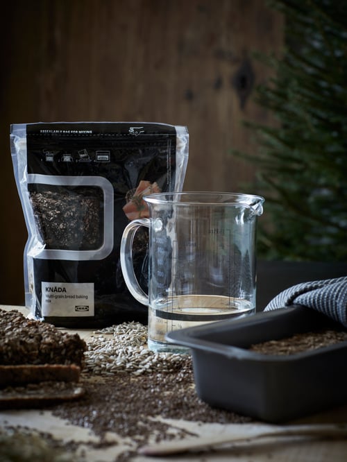 Auf einer Arbeitsplatte stehen eine Packung KNÅDA Backmischung Mehrkornbrot, Wasser in einem VARDAGEN Messbecher und Teig in einer HEMMABAK Brotform.