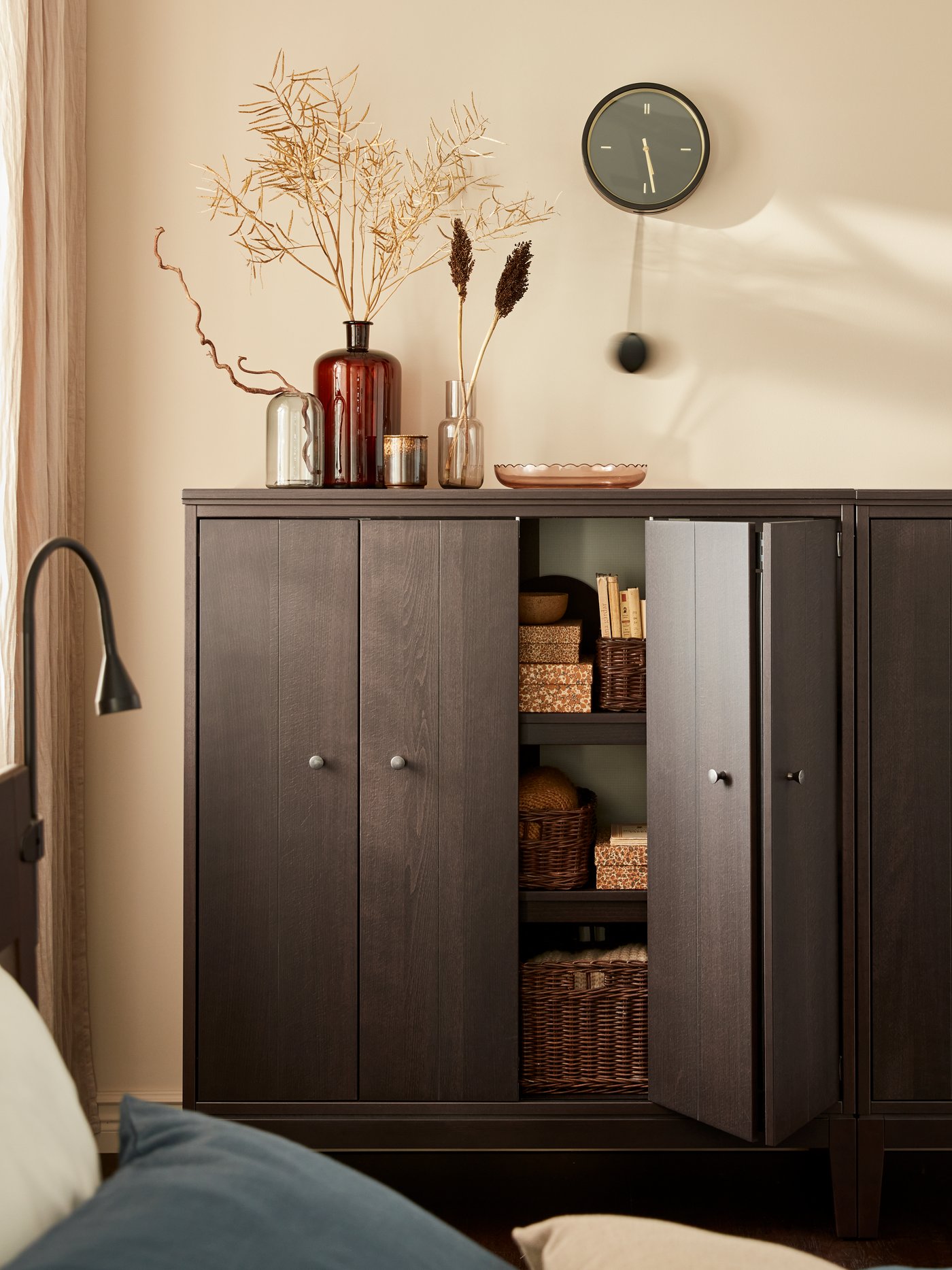 A wooden cabinet with bi-folding doors against a beige wall, with woven baskets and boxes in it. Vases and a clock.