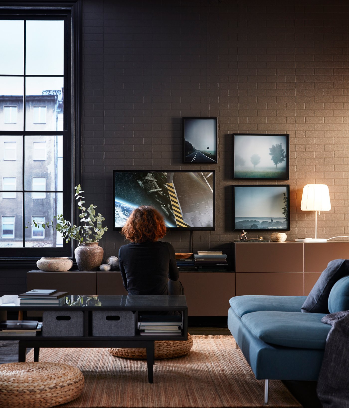 En person sitter på en sittpuff i ett vardagsrum och tittar på en TV. Runt TV:en hänger 3 små tavlor.