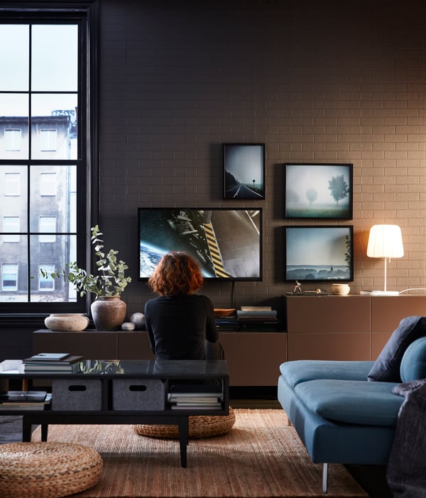 En person sitter på en sittpuff i ett vardagsrum och tittar på en TV. Runt TV:en hänger 3 små tavlor. 