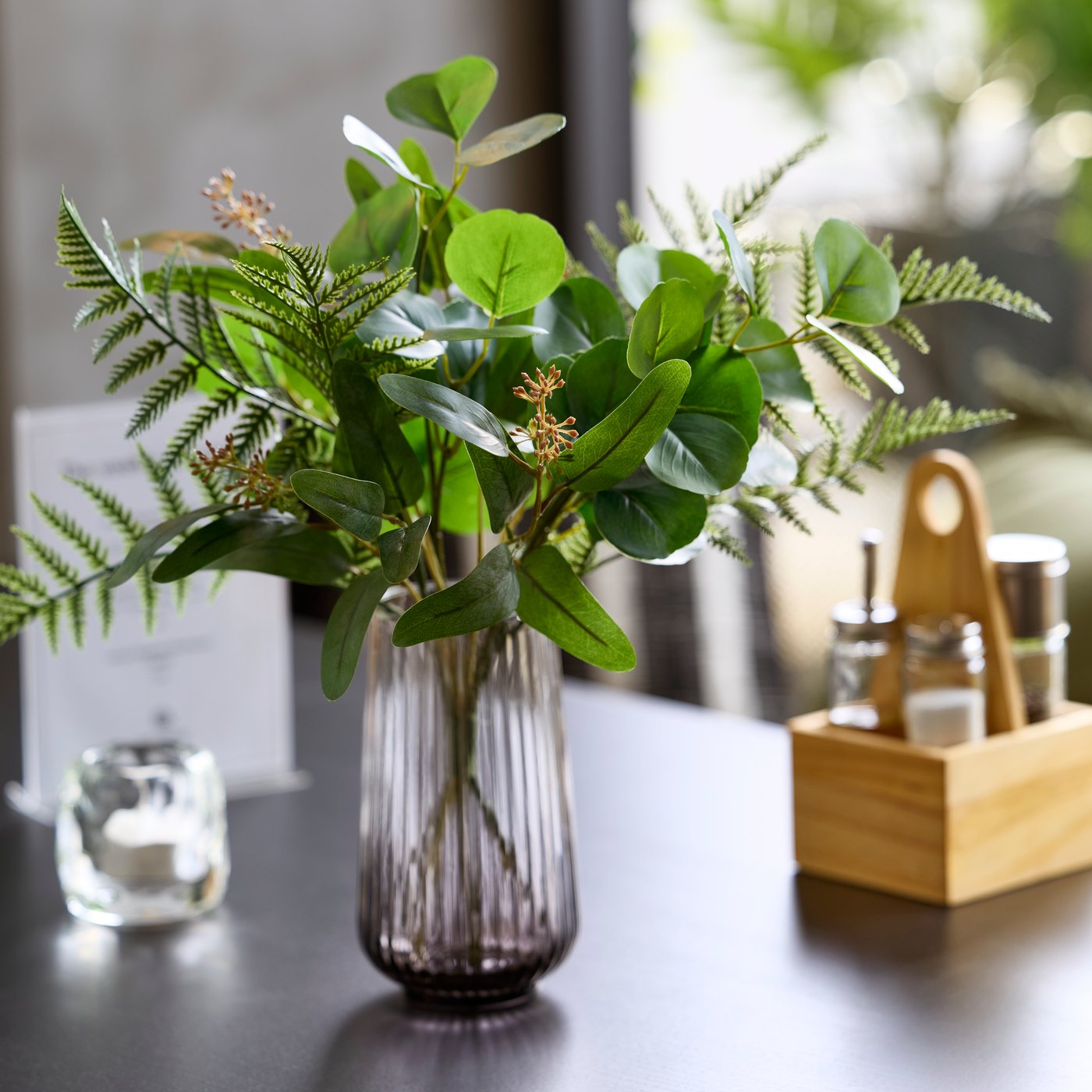 Close-up of a grey GRADVIS vase with greenery beside a wooden CHOKLADHAJ organiser and a LED tealight on a table.