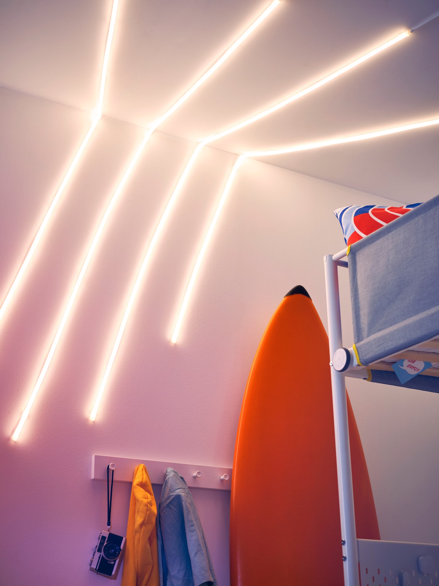 Ungdomsværelse med sejt lys af skinner på væggen. Surfbræt, skrivebord og kommode fylder værelset. 