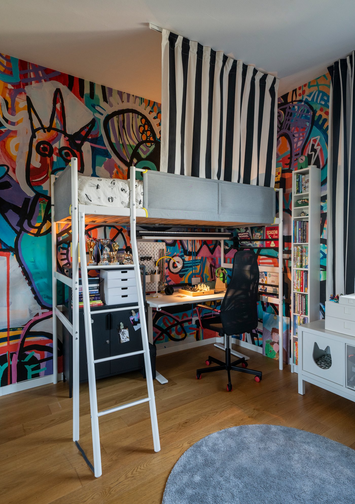 Cameretta colorata con letto a soppalco e tenda a righe bianche e nere, zona studio sotto il letto e pareti decorate con grandi murales grafici.