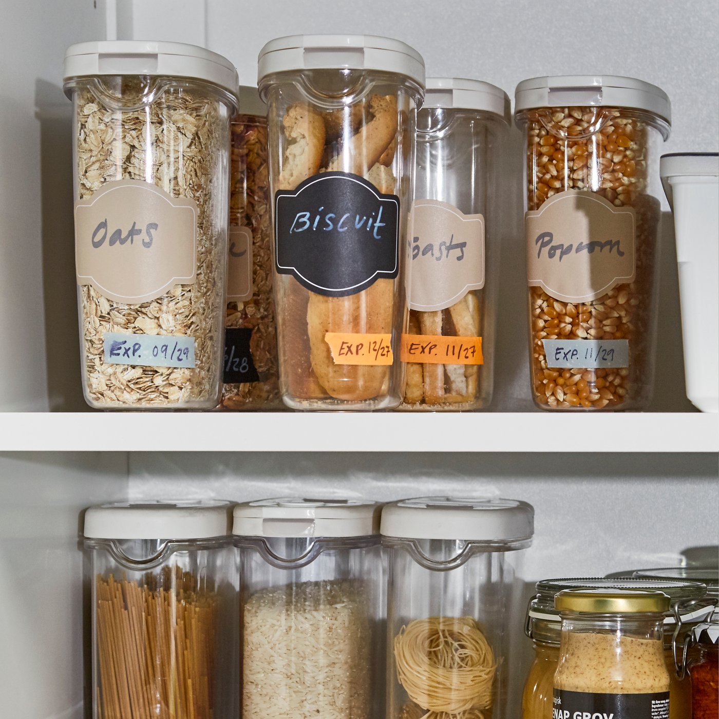 Different sized plastic food storage containers with labels populate two shelves in a kitchen cupboard.