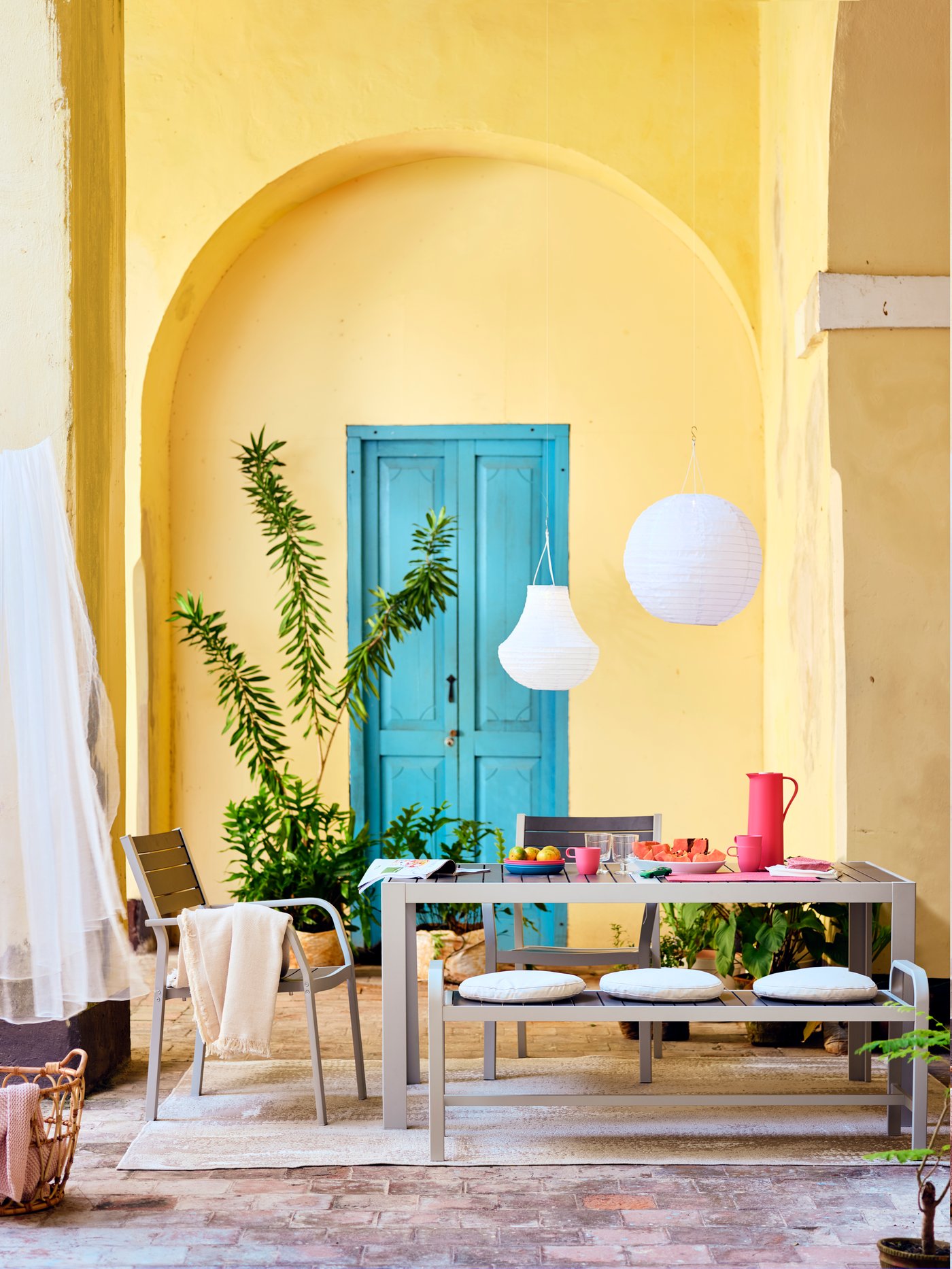 A white/beige TORPARÖ dining set with refreshments, a cutting board and a STORHAGA table lamp on top, and plants all around.