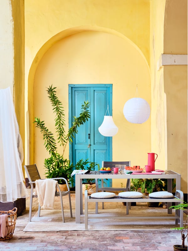 A white/beige TORPARÖ dining set with refreshments, a cutting board and a STORHAGA table lamp on top, and plants all around.