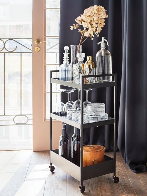 Une desserte à roulettes NISSAFORS en acier peint noir contient divers verres, des bouteilles et une plante dans un vase près d’une porte de balcon.