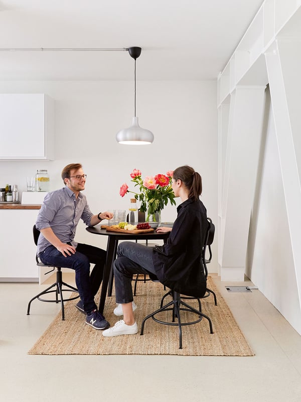 Deux personnes sont assises en riant à une table de salle à manger ronde dans la cuisine-café d'un cabinet d'avocats. Sur la table, il y a des snacks et un bouquet de fleurs dans un vase.