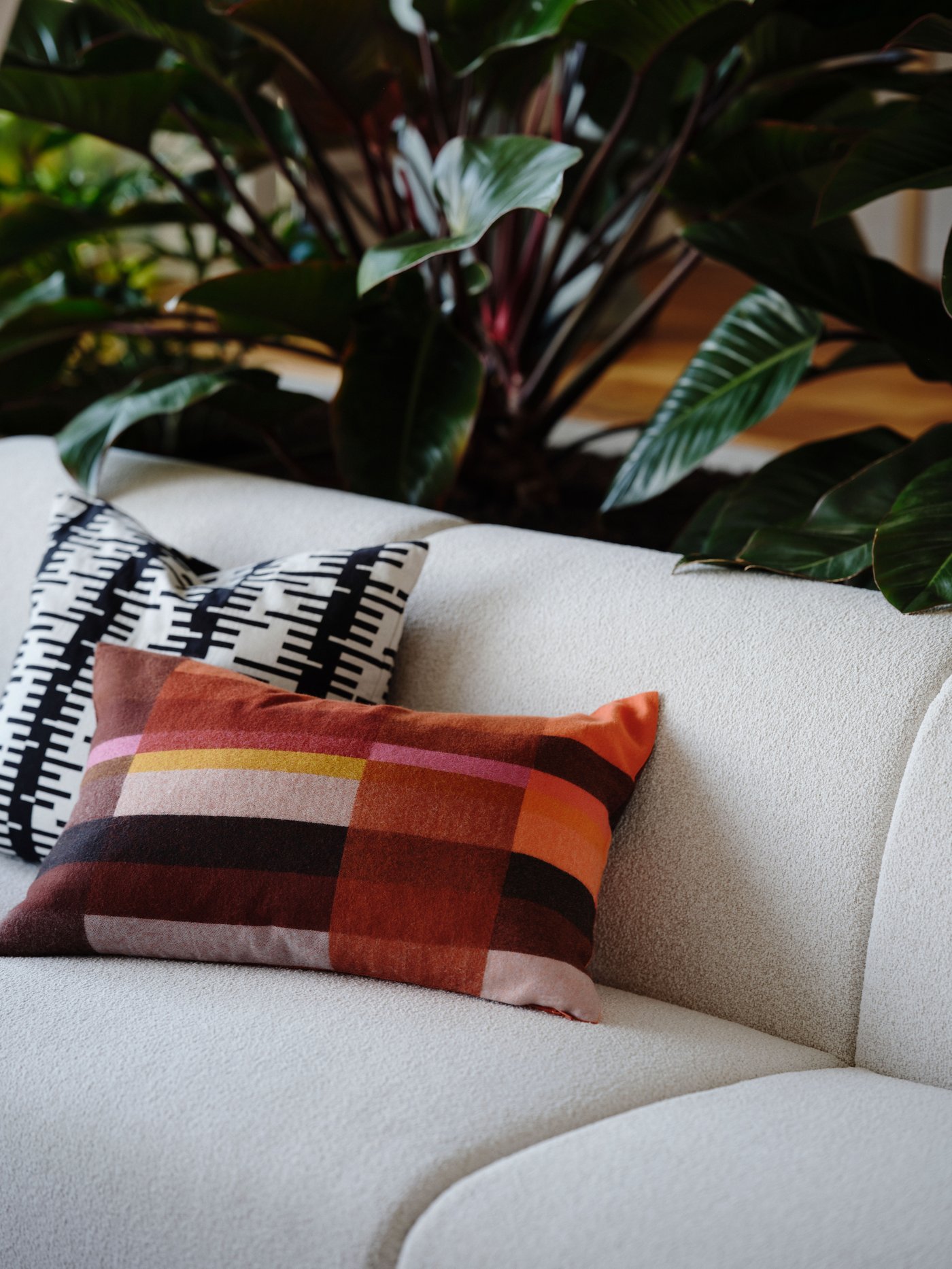 A sofa, with two cushions with different STOCKHOLM 2025 cushion covers on it, stands in front of some plants.