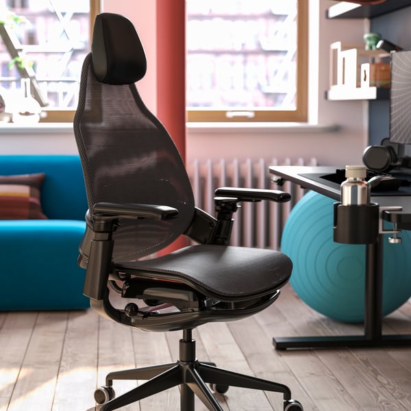 A black BÄSTBOLL gaming chair is beside a gaming desk in a bright room. A pillar and a blue yoga ball are near the chair.