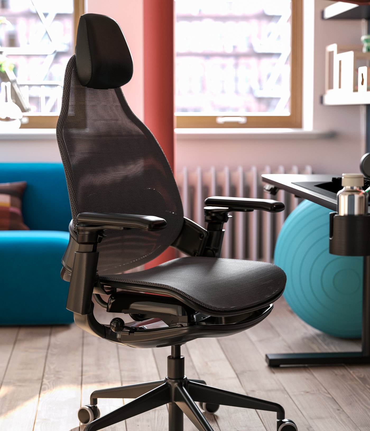 A black BÄSTBOLL gaming chair is beside a gaming desk in a bright room. A pillar and a blue yoga ball are near the chair.