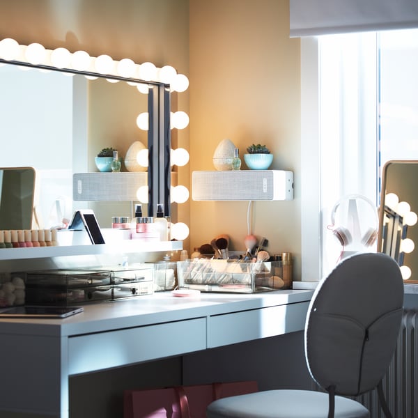 A big rectangular mirror with wall lamps above a white desk with lots of make-up storage, plus a white speaker on the wall.