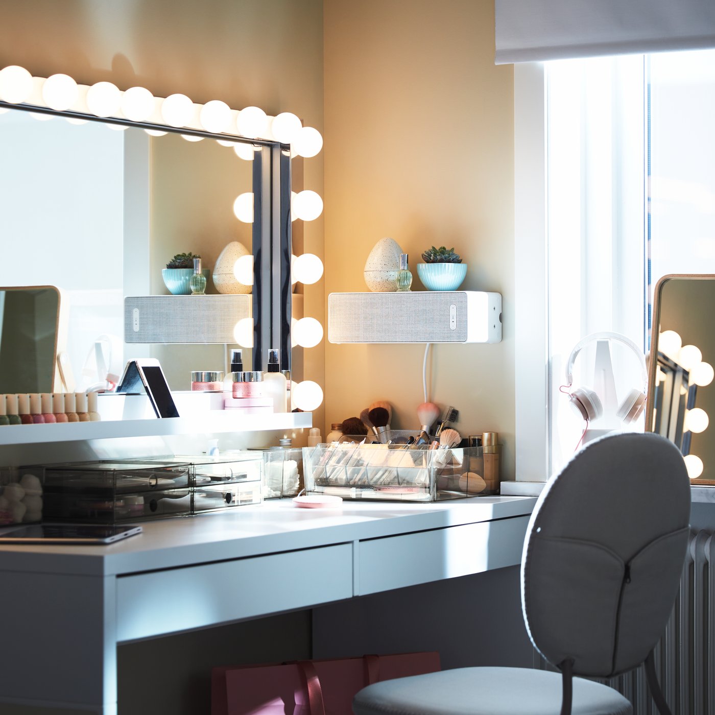 A big rectangular mirror with wall lamps above a white desk with lots of make-up storage, plus a white speaker on the wall.