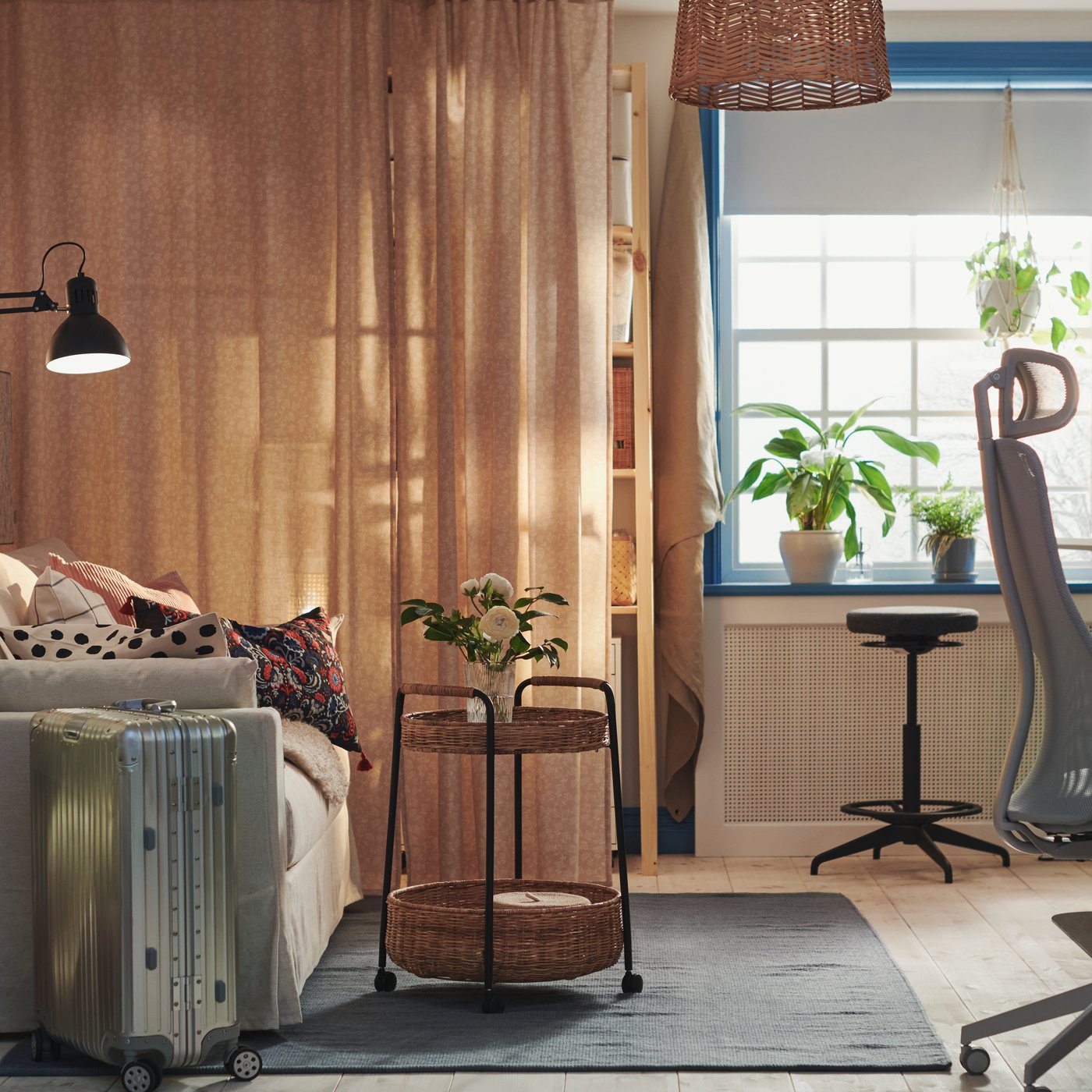 Una stanza con un carrello con vano contenitore su un tappeto, davanti a un divano letto HYLTARP e vicino a uno sgabello per seduta attiva LIDKULLEN. Sul lato destro della stanza si vede una scrivania con una sedia da gaming, una cassettiera, una tastiera, delle cuffie, un mouse e un monitor. Sul fondo della stanza c'è una finestra sul cui davanzale ci sono due piante e sotto uno sgabello. 