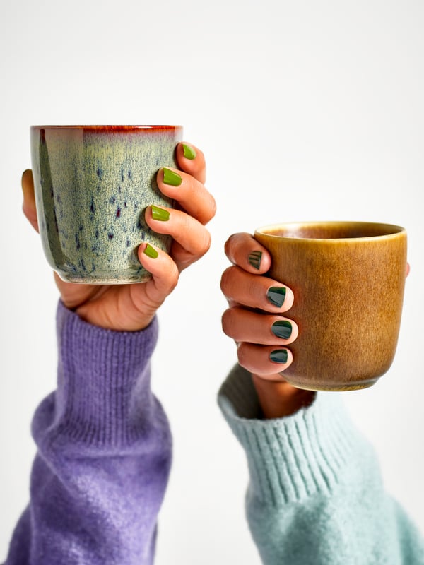 Two hands close to each other are holding two GLADELIG mugs in mixed colours against a white background.
