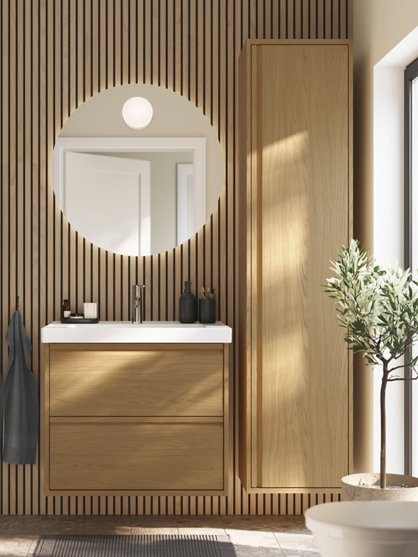 A bathroom with wooden walls and a window has an ÄNGSJÖN wash-stand and a wall cabinet in oak effect and a rug on the floor.