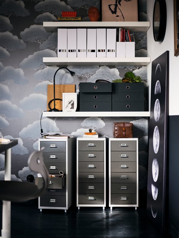 Beneath three white LACK shelves mounted on an artfully patterned wall, stand three side-by-side grey HELMER drawer units.