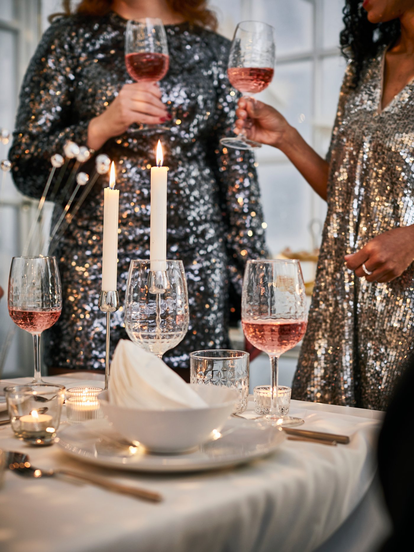 Two people in sparkly dresses raise their wine glasses to toast the New Year. The table is festively set with white crockery and gold cutlery.