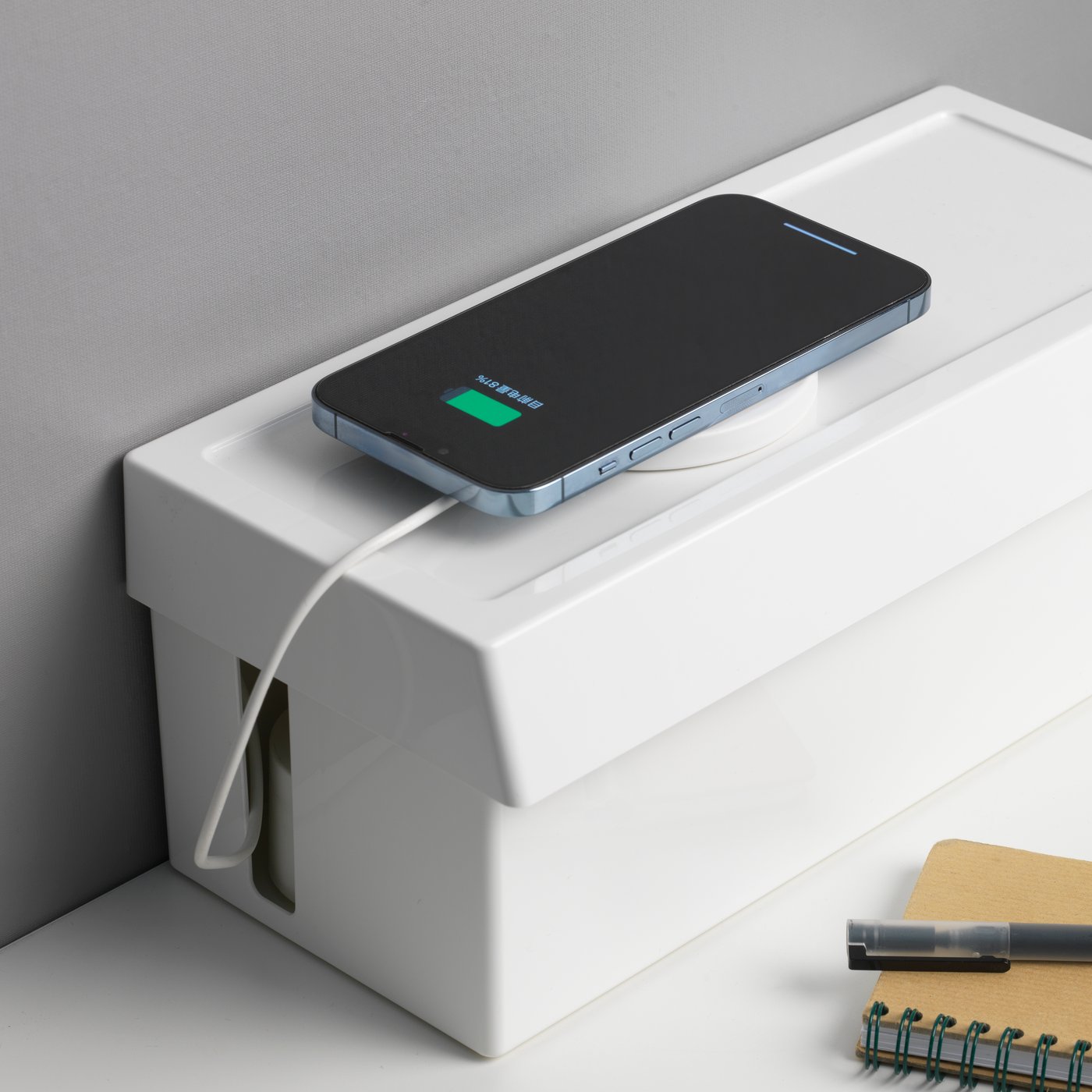 Uno smartphone in carica la cui presa si trova in una scatola per cavi con coperchio SÄTTING. Accanto, un'agenda con una penna.