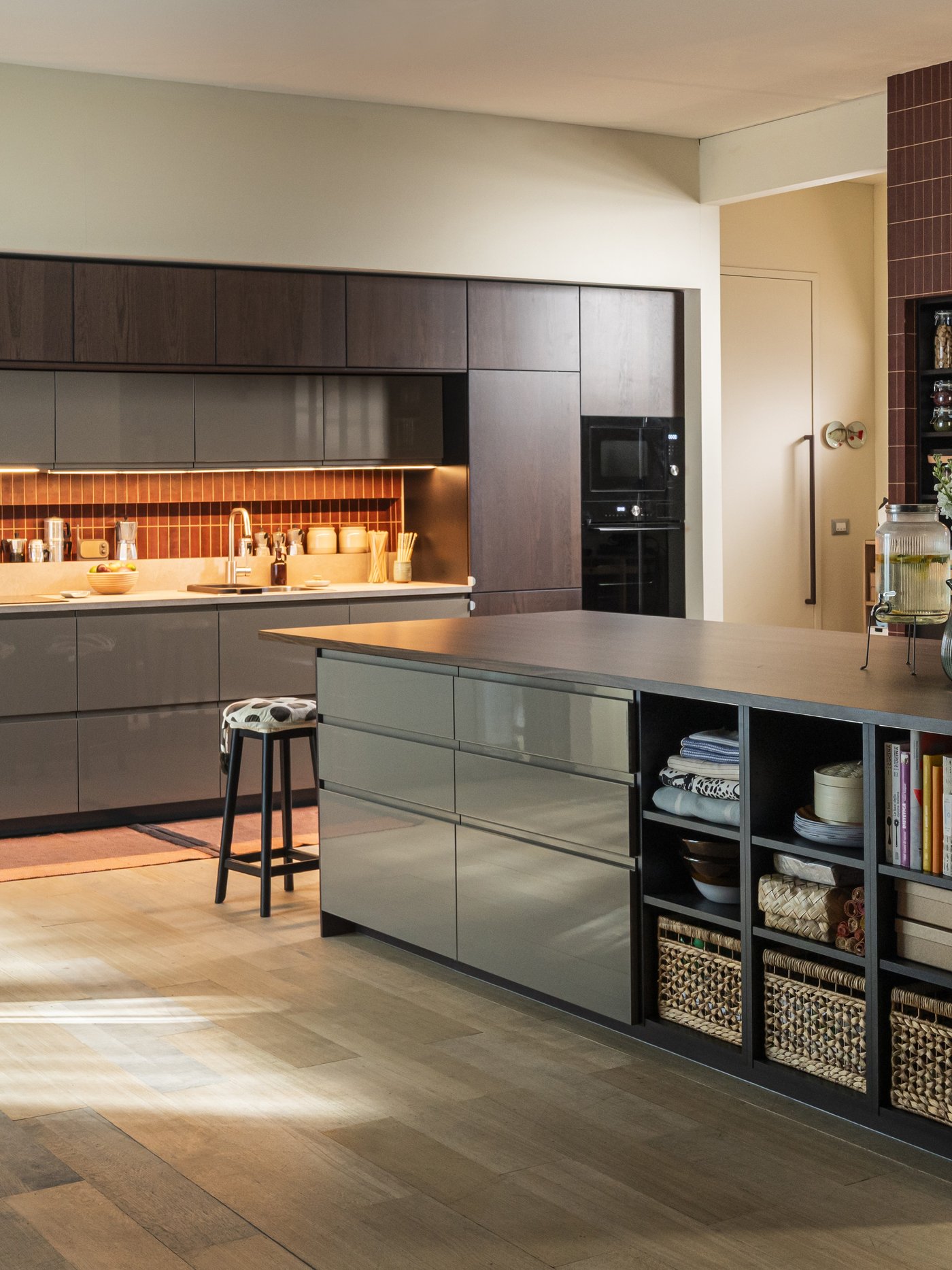 Cucina moderna IKEA con isola centrale, finiture in legno scuro, ampi piani di lavoro e dettagli organizzativi come vassoi, barattoli e cesti negli scaffali. Sullo sfondo parete attrezzata e scaffali con ante a vetro.