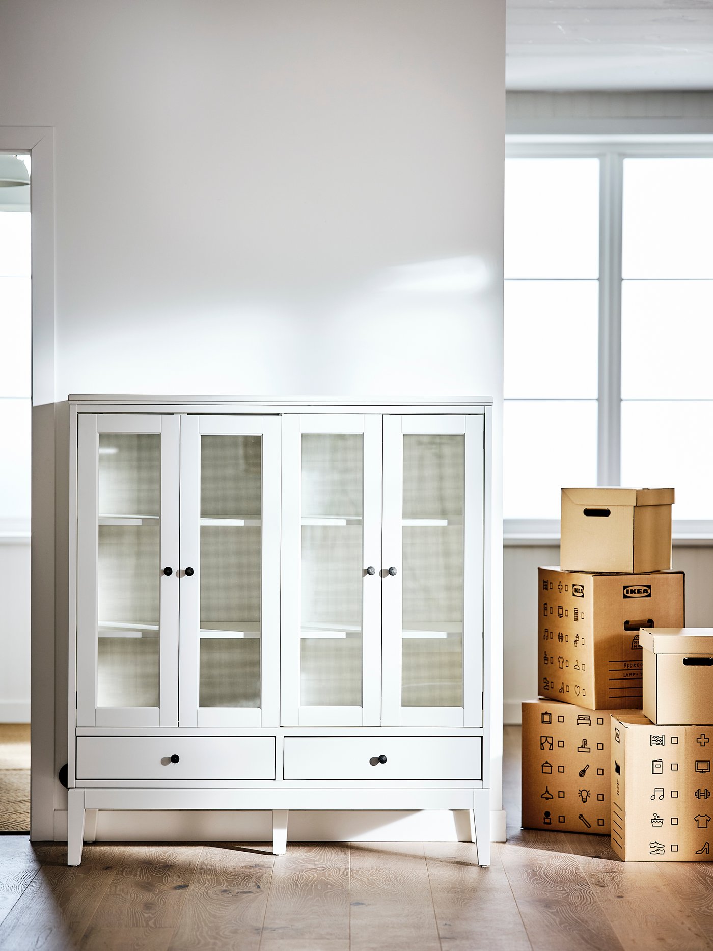 Living con un armario blanco con puertas de vidrio plegables con estantes regulables y patas, junto a 5 cajas.